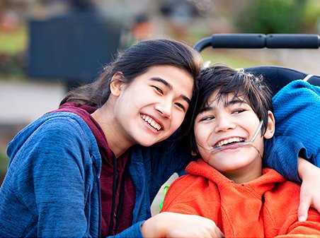 Big sister hugging disabled brother in wheelchair outdoors, smiling