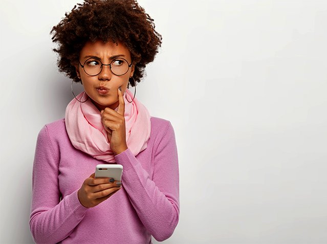 Pensive dark skinned woman looks thoughtfully aside, holds mobile phone, waits for call, purses lips, has Afro haircut, dressed in violet jumper with scarf, keeps index finger on cheek, poses indoor Pensive dark skinned woman looks thoughtfully aside, holds mobile phone, waits for call, purses lips, has Afro haircut, dressed in violet jumper with scarf, keeps index finger on cheek, poses indoor